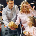 A family enjoying shopping in a supermarket aisle, selecting groceries with a cart.